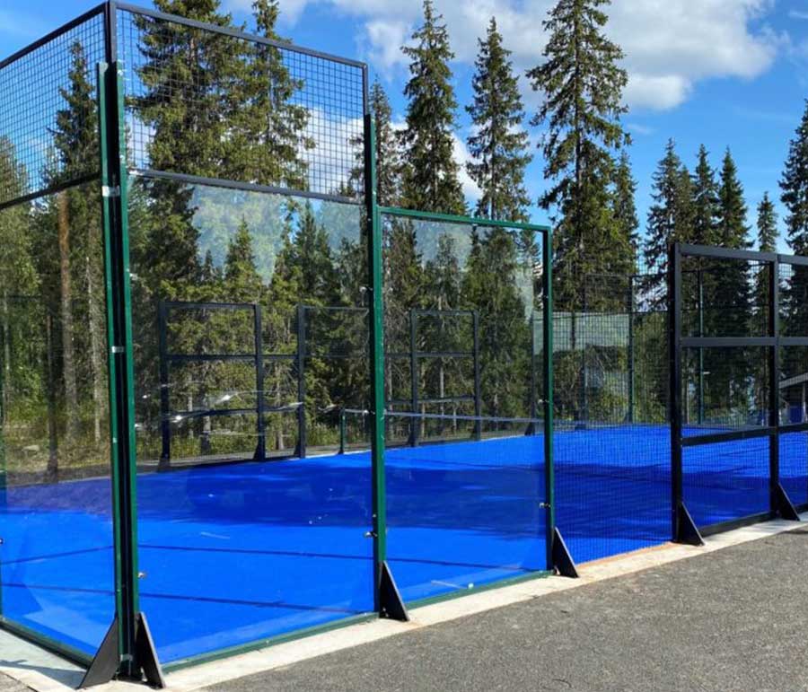 Outdoor glass-walled padel court with a blue playing surface, surrounded by tall pine trees under a clear blue sky at Sameland Retreat in Swedish Lapland.