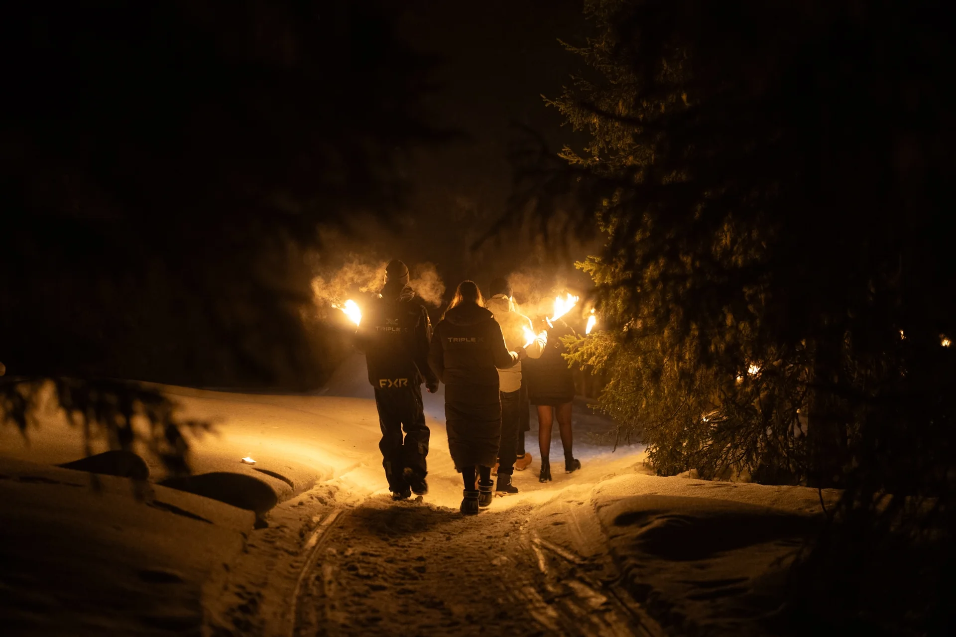 Guests walking a snowy forest path at night holding burning torches that light the trees.